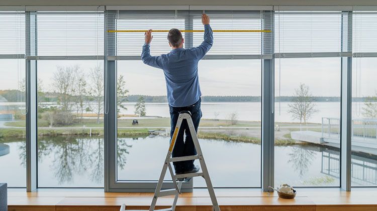 A man in a room on ladder is showing how to measure for window blinds